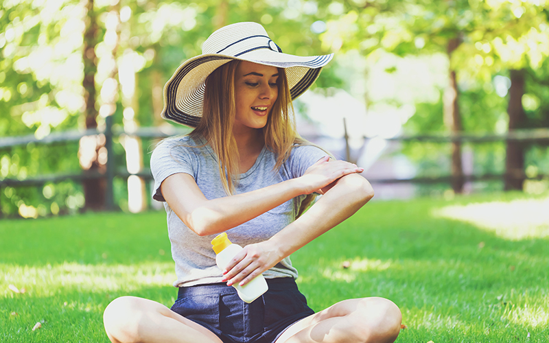 Young woman wearing a sun hat, sitting on the grass applying sunscreen